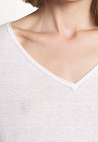 Nahaufnahme eines Halses und Schlüsselbeins einer Person, die ein weißes V-Ausschnitt-Shirt trägt, mit kurz geschnittenem braunen Haar, das teilweise sichtbar ist.