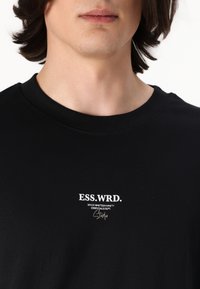 Fekete pamut póló, kerek nyakkivágással és fehér logó felirattal az elején, amely a "ESS.WRD." és a "SINCE NINETEEN NINETY." szöveget tartalmazza.