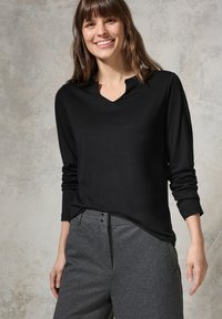 Maglietta nera a maniche lunghe con scollo a V e polsini arrotolati, abbinata a pantaloni grigi testurizzati con dettagli a bottoni.