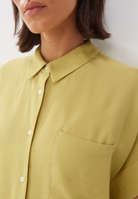 Camicia gialla con colletto, realizzata in tessuto morbido e texturizzato, caratterizzata da una chiusura con bottoni e un'unica tasca sul petto.