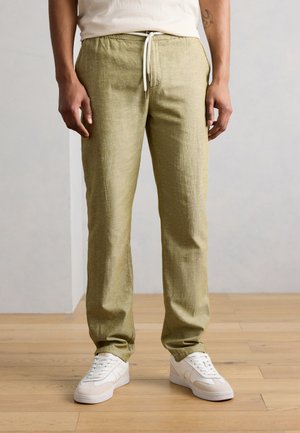 Άνδρας που φοράει ανοιχτό καφέ παντελόνι με κορδόνι και λευκά αθλητικά παπούτσια, στέκεται σε ξύλινο πάτωμα μπροστά σε γκρι τοίχο.