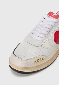 Hvit skinnsneaker med perforert tå, rød logo aksent og gullbokstaver. Har en teksturert overflate og en gummisål.