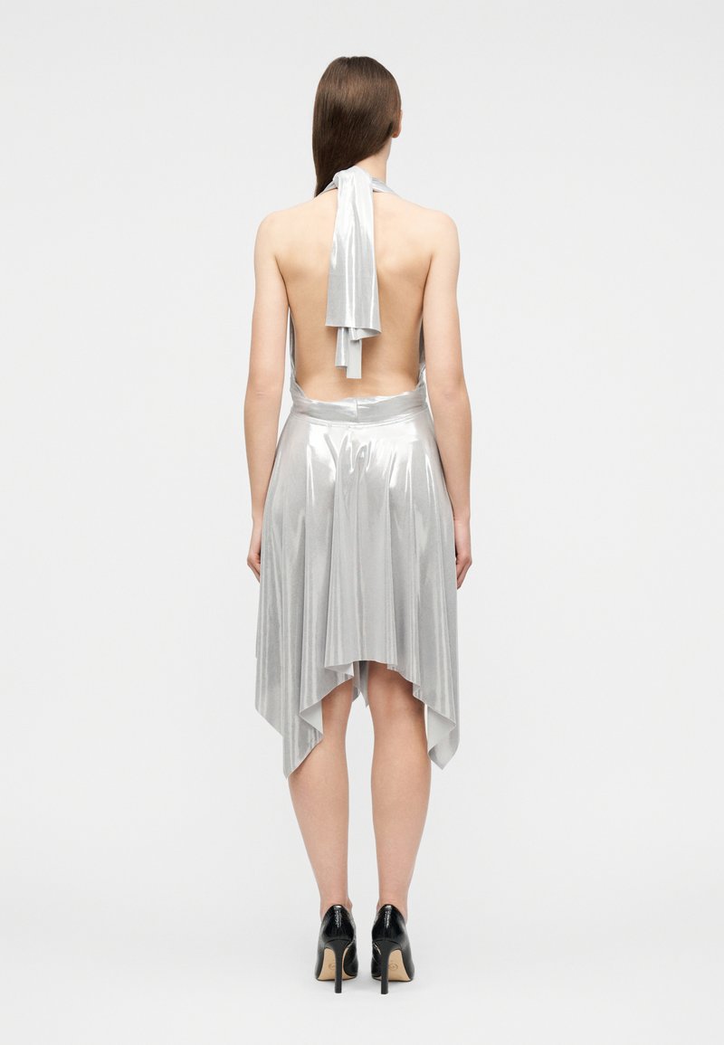 Femme portant une robe dos nu argentée à col halter avec un ourlet asymétrique, associée à des chaussures à talons hauts noires, debout, dos tourné.