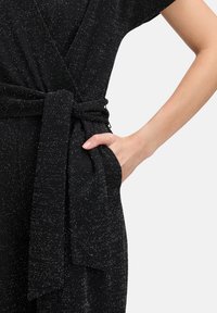 Robe noire à encolure cache-cœur avec une ceinture à nouer à la taille, manches courtes et tissu texturé aux paillettes métalliques. Une main repose dans une poche latérale.