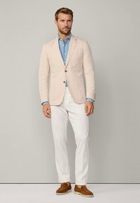 Blazer de linho bege claro por cima de uma camisa azul clara com botões, combinado com calças brancas e sapatos de camurça castanhos. Design simples e ajustado.