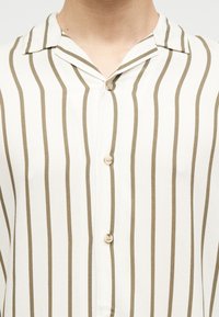 Λευκό πουκάμισο με κάθετες ρίγες σε ελιά πράσινο, ανοιχτός γιακάς και τρία ορατά κουμπιά στο μπροστινό μέρος.