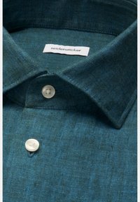 Gros plan du col d'une chemise habillée verte texturée avec deux boutons blancs et étiquette de la marque "seidensticker" à l'intérieur.