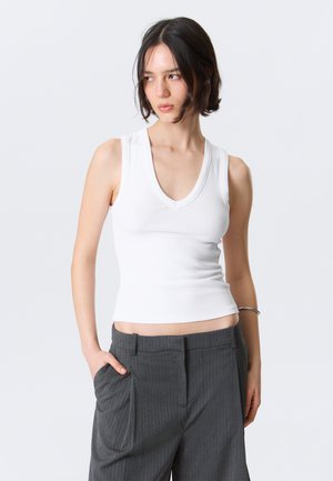 Jeune personne aux cheveux courts et foncés portant un haut blanc sans manches à côtes et un pantalon gris à fines rayures, debout avec une main dans la poche.