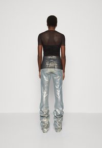 Top negro ajustado y transparente con detalles fruncidos, combinado con jeans azules metálicos de diseño holgado y textura brillante, visto desde la parte de atrás.