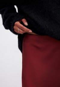 Schwarzer, flauschiger Pullover mit langen Ärmeln, kombiniert mit einem glatten burgunderfarbenen Satrock. Hand sichtbar, die den Rand des Pullovers hält, silberner Ring an einem Finger.