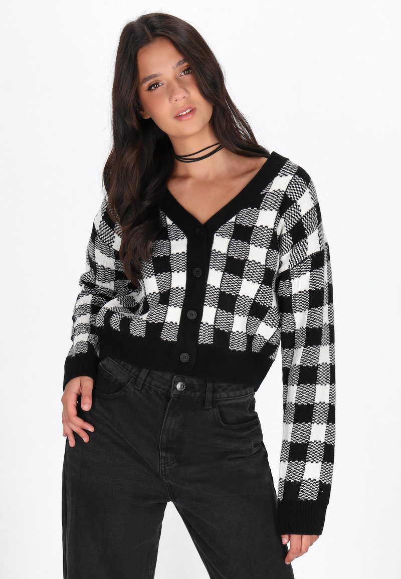 Cardigan a scacchi bianco e nero con scollo a V, maniche lunghe e bottoni lungo la parte frontale. Indossato sopra jeans neri a vita alta.