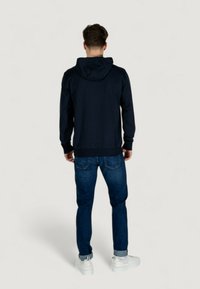 Sweatshirt à capuche bleu marine avec manches longues, poignets et ourlet côtelés, associé à un jean en denim sombre et des baskets blanches. Tissu texturé, design simple.