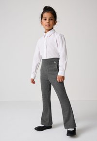 Junges Mädchen steht und trägt ein weißes Hemd mit Knöpfen, graue ausgestellte Hosen, weiße Socken und schwarze Schuhe vor einem einfarbigen Hintergrund.