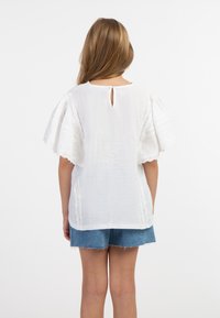 Hvid bomuldsbluse med flagrende ærmer, med broderede detaljer langs kanterne og en nøglehulåbning bagpå. Parret med denimshorts.