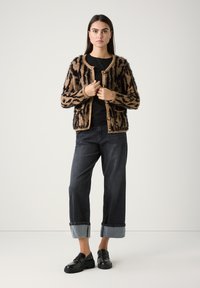 Flauschige cardigan in Braun und Schwarz mit Muster, Rundhalsausschnitt und Knopfverschluss, getragen über einem schwarzen Top, kombiniert mit dunklen Jeans und schwarzen Slippern.
