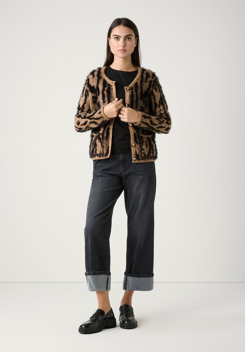 Flauschige cardigan in Braun und Schwarz mit Muster, Rundhalsausschnitt und Knopfverschluss, getragen über einem schwarzen Top, kombiniert mit dunklen Jeans und schwarzen Slippern.