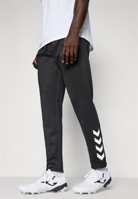 Mann iført svarte treningbukser med hvite chevronstriper på leggene, hvite sportssko og en hvit skjorte, står mot en grå bakgrunn.