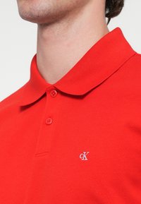 Piros anyagú texturált póló, rövid ujjú, klasszikus gallérral. Két gombbal és egy kis hímzett logóval a mellkasán.