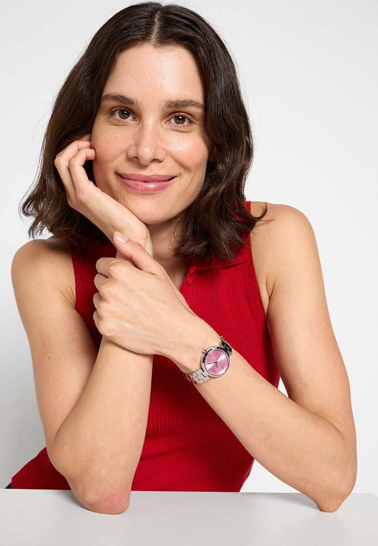 Montre argentée avec un cadran rose rond, bracelet en métal à maillons et marqueurs horaires simples. Le modèle porte un haut rouge côtelé sans manches, les mains jointes.