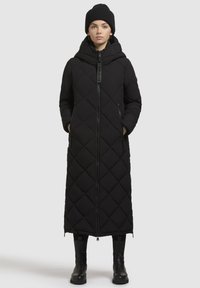 Schwarzer, gesteppter langer Mantel mit Kapuze, ausgestattet mit einem Reißverschluss auf der Vorderseite, Seitentaschen und einem Rautenmuster. Kombiniert mit einer schwarzen Strickmütze und Stiefeln.