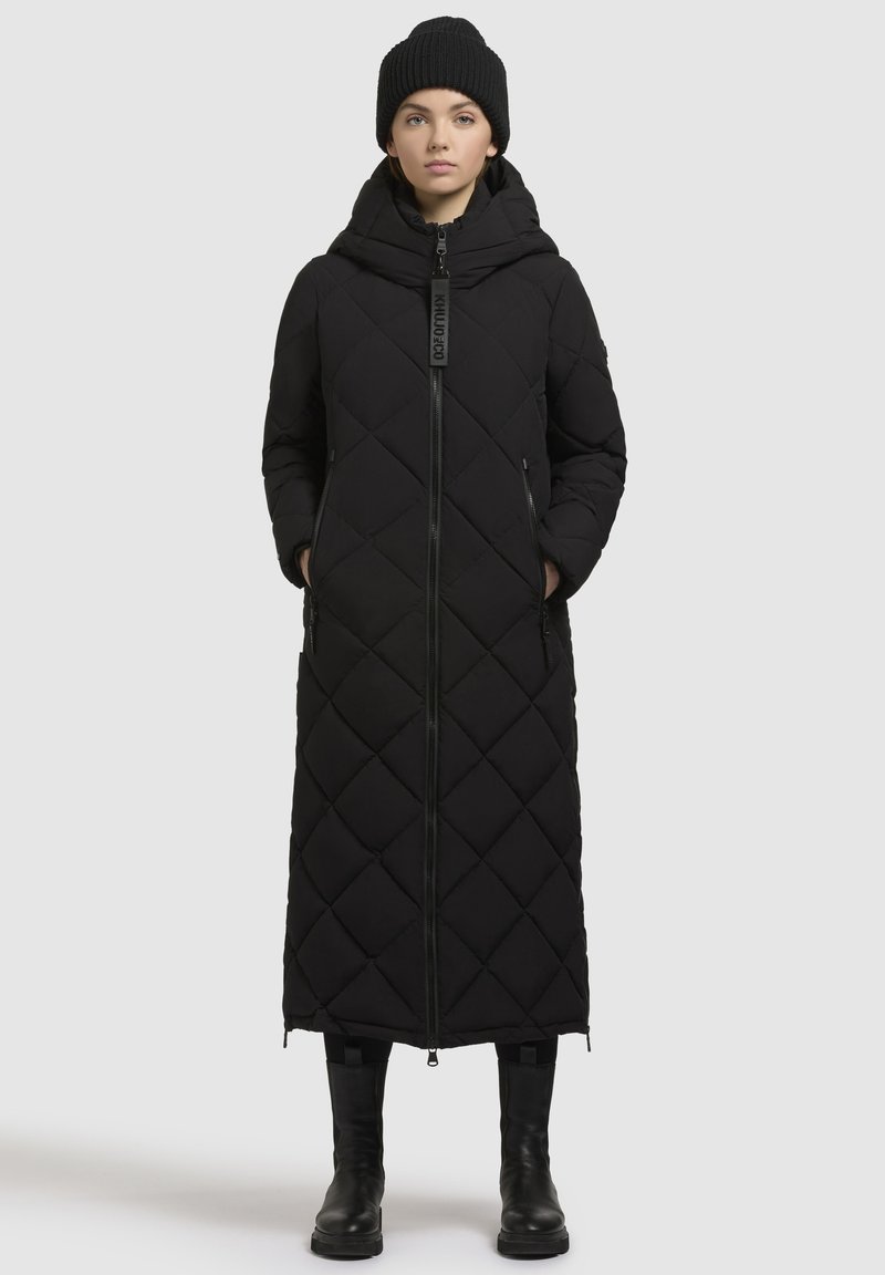 Schwarzer, gesteppter langer Mantel mit Kapuze, ausgestattet mit einem Reißverschluss auf der Vorderseite, Seitentaschen und einem Rautenmuster. Kombiniert mit einer schwarzen Strickmütze und Stiefeln.