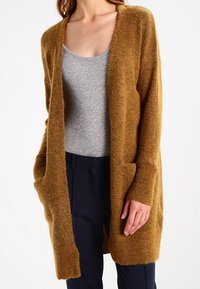 Cárdigan largo de apertura frontal en color mostaza, hecho de un material suave y peludo. Presenta dos bolsillos laterales y puños de canalé.