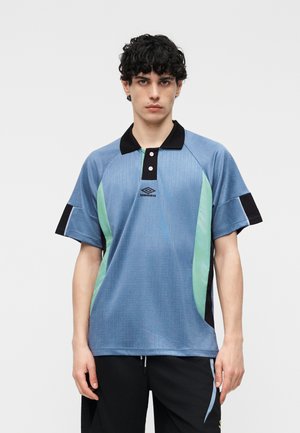 Jonge man met krullend zwart haar, gekleed in een blauw-groen polohemd met korte mouwen, een zwarte kraag en zwarte broek.