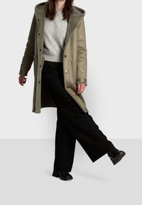 Cappotto lungo color verde oliva con cappuccio, dotato di una morbida fodera interna, chiusura con bottoni e tasche laterali. Abbinato a pantaloni neri e stivali.