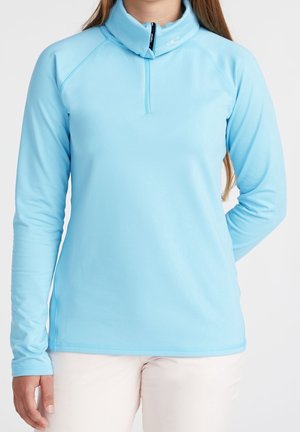 Person trägt einen hellblauen Langarm-Quarter-Zip-Pullover mit Stehkragen und weiße Hose, steht vor einem einfarbigen Hintergrund.