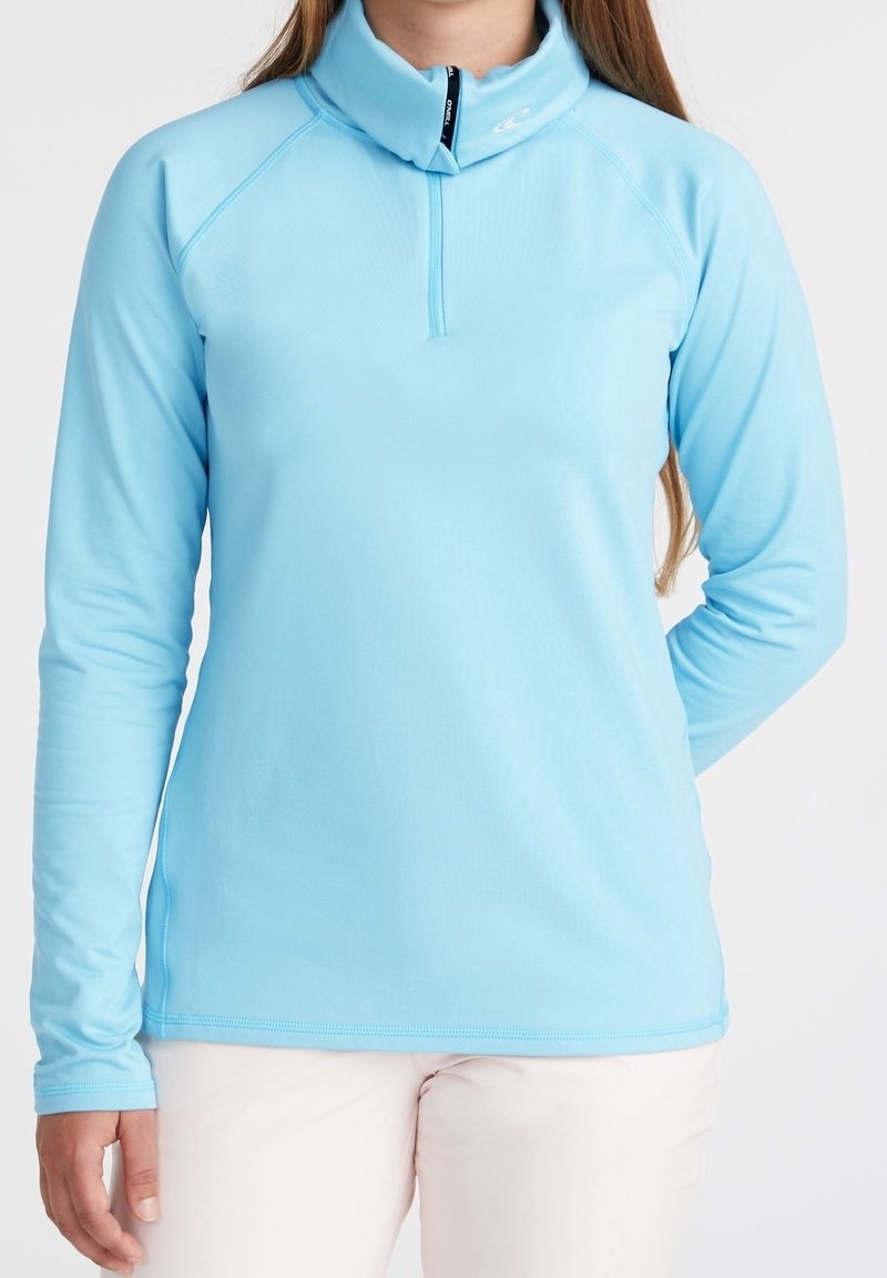 Personne portant un pull à demi-zip bleu clair à manches longues avec un col montant et un pantalon blanc, debout devant un fond uni.