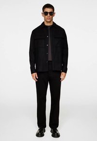 Schwarze Hemdjacke mit zwei Brusttaschen, Knopfleiste vorne und lässiger Passform. Kombiniert mit schwarzen Hosen und klobigen schwarzen Schuhen. Sonnenbrille getragen.