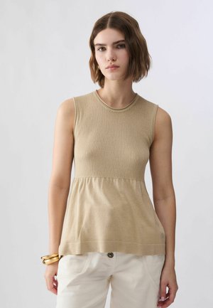 Femme aux cheveux courts bruns portant un haut beige sans manches et un pantalon blanc, debout face à l’avant, les bras détendus et une expression neutre.