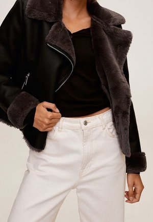 Person iført en sort jakke med mørk pelsforing og manchetter, en sort top og hvide højtaljede jeans med knap og lynlås.
