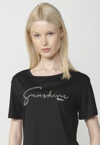 Camiseta negra de manga corta en tejido suave, con texto de brillantes rhinestones plateados. Diseño de cuello redondo con corte relajado y textura suave.