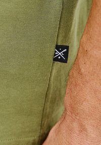 Oliwkowa bawełniana koszulka z gładką fakturą. Posiada czarną metkę z białym logo skrzyżowanych igły i widelca na dolnym brzegu.