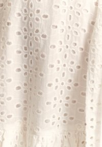 Bílá látka s vyšívaným žakárovým vzorem, obsahující kruhové otvory a květinové tvary, s texturou roztřepených okrajů na lemu.
