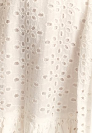 Witte stof met geborduurd oogje-patroon, met cirkelvormige openingen en bloemmotieven, getextureerd met rafelige randen aan de zoom.