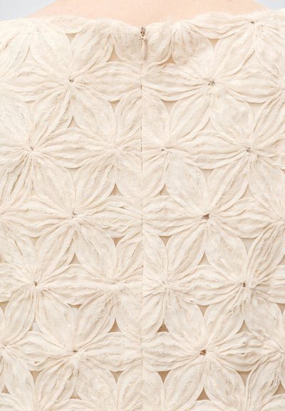 Tissu crème avec un motif floral texturé composé de formes de pétales superposées et une fermeture éclair centrale en haut.