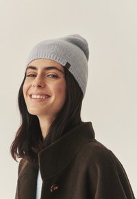 Hele helesinise kootud beanie, millel on volditud serv ja väike pruun silt. Kootud tumepruuni mantliga, neutraalse taustaga.