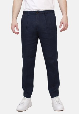 Uomo che indossa pantaloni plissettati blu scuro con vita abbottonata e sneakers bianche, in piedi con le braccia rilassate ai lati.