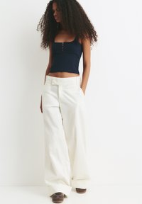 Vrouw met lang krullend haar die een marineblauwe crop top en brede witte broeken draagt, met handen in de zakken, en bruine sneakers voor een effen witte achtergrond.