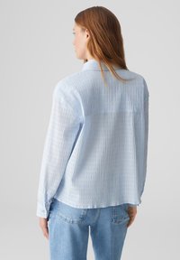 Camicia azzurro chiaro con un design a pieghe e texture, maniche lunghe e un dettaglio sullo yoke posteriore, abbinata a jeans in denim blu.
