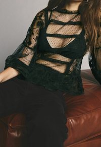 Sort gennemsigtig bluse med prikmønster, blomsterlacemærker og flæsevedstående. Har et lagdelt design med lange klokkeærmer.