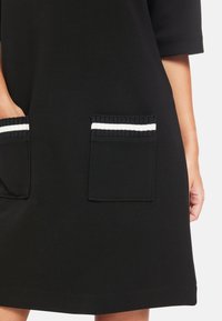 Schwarzes Kleid mit kurzen Ärmeln, zwei vorderen Taschen mit strukturiertem dunklem Ripp und weißen Streifen an den Taschenkanten. Glatte Stofftextur.