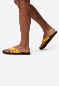Sorte og gule flip-flops med en tekstureret sål, der har en blød stofstrop og brandlogoet på den øverste strop.