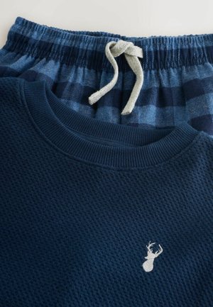 Námornícka modrá mikina s vaflovou štruktúrou a malým logom jeleňa, spárovaná s modrými kockovanými pyžamovými nohavicami s elastickým pásom a šnúrkou.