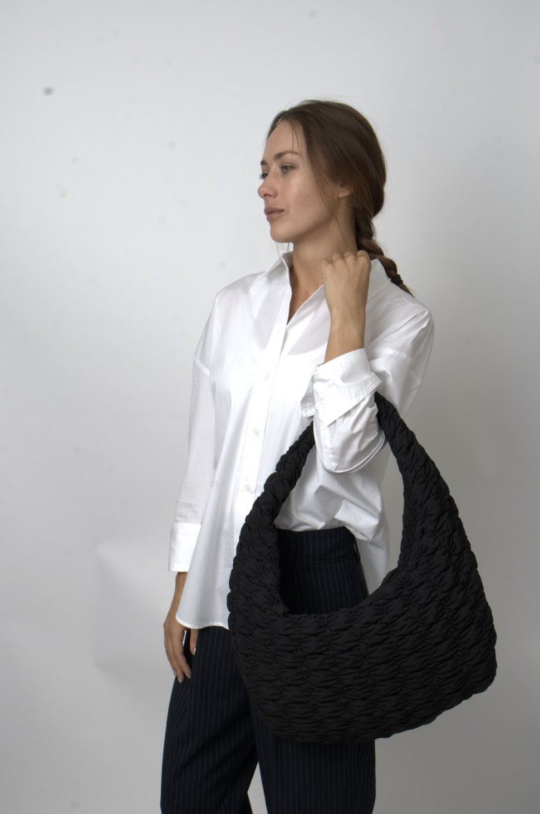 Mulher em camisa branca e calças com riscas a segurar uma grande mala de ombro preta texturizada contra um fundo simples.