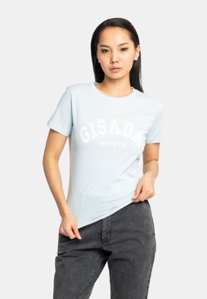 Femme aux longs cheveux foncés portant un t-shirt bleu clair Gisada Switzerland et un pantalon gris foncé, ajustant son t-shirt sur un fond blanc.