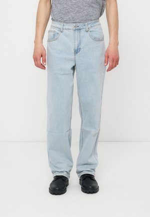 Man met lichtblauwe straight-leg jeans, grijs gestreept overhemd in de broek gestopt, en zwarte veterschoenen, staand tegen een witte achtergrond.