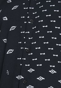 Svart kortärmad skjorta med vit rosett och geometriska mönster. Har knappar och en mjuk tygtextur.